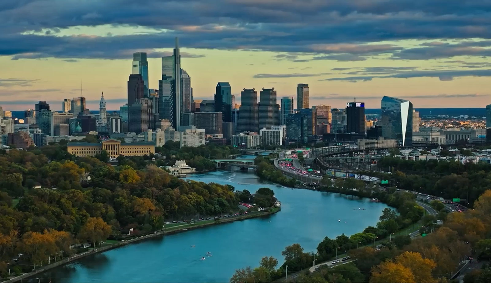 Aerial view of a city skyline at dusk with skyscrapers under a cloudy sky. A river with rowing boats flows through lush green trees in the foreground.